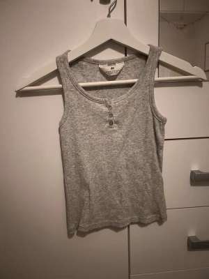 Grå ribbad linnetopp från H&M - Säljer en enkel och snygg grå ribbad linnetopp från H&M. Toppen har rund halsringning och tre små knappar framtill för en chill look. Perfekt till jeans eller shorts under varmare dagar.