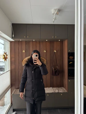 Svart dunjacka med pälskrage från Hollies - Varm och skön svart quiltad dunjacka från Hollies med stor fluffig avtagbar pälskrage. Jackan har dragkedja, knappar, två fickor framtill och innerficka. Mycket gott skick. 