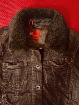 Brun manchesterjacka med fuskpäls från Esprit M - Snygg brun manchesterjacka från Esprit med bred krage i fuskpäls. Jackan har två bröstfickor med knapp, dekorativa sömmar och knäppning framtill. Insidan har ett mönstrat foder och coola detaljer med röd etikett och snöre.