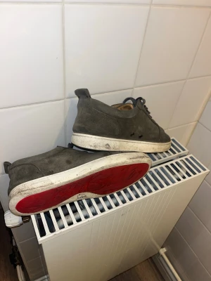 Grå mockasneakers från Christian Louboutin - Säljer ett par grå sneakers från Christian Louboutin i mocka med klassisk röd sula och vita kanter. Skorna har rund tå, svarta snören och diskreta metalldetaljer vid snörningen. Insidan är beige och materialet känns lyxigt och mjukt.