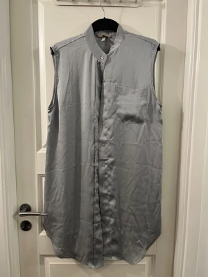 Grå ärmlös blus från H&M - Säljer en stilren grå ärmlös blus från H&M i storlek M. Blusen har klassisk krage, knäppning framtill och en bröstficka. Materialet är mjukt och har en lätt glansig finish som ger en snygg look.