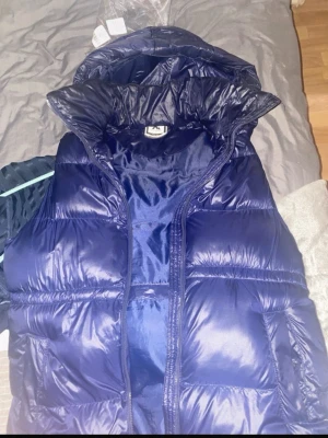 Mörkblå dunväst med huva - Säljer en mörkblå dunväst med hög krage och huva. Västen har en glansig finish och stängs med dragkedja framtill. Perfekt att slänga över en hoodie för extra värme. Fylld med dun och har två fickor på sidorna.