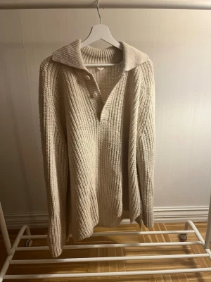 Beige stickad tröja med krage - Säljer en beige stickad tröja med bred krage och knappar vid halsen. Tröjan har en ribbad struktur och långa ärmar, perfekt för kyliga dagar. Passformen är loose och materialet känns mjukt och mysigt. Storlek M men passar som L!