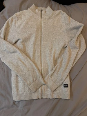 Ljusgrå kofta från Jack & Jones - Säljer en stilren ljusgrå kofta från Jack & Jones med hel dragkedja framtill och ribbade muddar. Perfekt till lager på lager och enkel att matcha med olika outfits. Mjuk och skön känsla tack vare bomullsmaterialet.