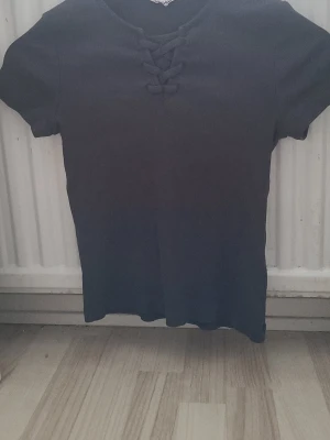 Svart ribbad topp med snörning - Säljer en svart ribbad topp med korta ärmar och snygg snörningsdetalj vid halsen. Toppen har en enkel, stilren design och är gjord i ett mjukt, stretchigt material som sitter bekvämt. Perfekt att matcha med jeans eller kjol.