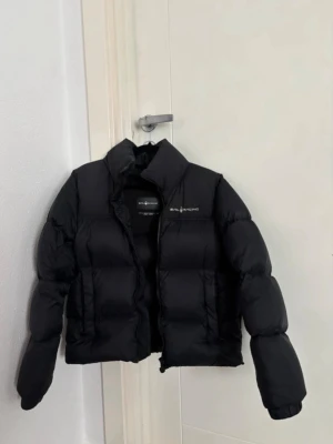 Svart pufferjacka från Sail Racing - Pris kan diskuteras! Svart pufferjacka från Sail Racing med hög krage och diskret logga på bröstet. Jackan har en quiltad design, dragkedja framtill och två sidofickor. Materialet är syntet och jackan har långärmade armar. Perfekt för kalla dagar och har en clean, modern look. 