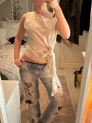 Beige omlottopp med knyt från Zara - Säljer en beige omlottopp från Zara i storlek M. Toppen har en cool asymmetrisk halsringning och knyt i midjan som ger en snygg siluett. Ärmlös design och mjukt bomullsmaterial gör den både bekväm och trendig. Perfekt att styla med jeans eller kjol🫶🏼