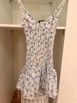 Blommig kortklänning med volang - Supersöt kortklänning med smala axelband och v-ringad hals. Klänningen är ljus med blått blommönster och har en volang nedtill. Smockad överdel ger en snygg passform och den är perfekt för varma dagar.