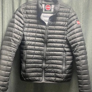 Svart pufferjacka från Colmar - Snygg svart pufferjacka från Colmar med quiltad design och diskret logga på ärmen. Jackan har dragkedja framtill, två sidofickor med dragkedja och en innerficka. Perfekt för dig som gillar stilren och sportig look.