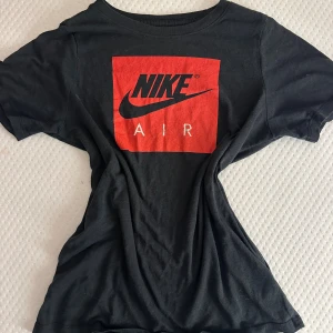 Svart Nike Air t-shirt med röd logga - Svart t-shirt från Nike med klassisk Air-logga i rött och svart framtill. T-shirten har rund halsringning och korta ärmar. Tillverkad i mjuk bomull för en skön känsla. Perfekt för dig som gillar sportig och stilren streetwear.
