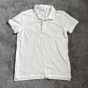 Vit pikétröja från Kappahl - Klassisk vit pikétröja från Kappahl i storlek 158/164. Tröjan har korta ärmar, krage och två knappar framtill. Tillverkad i mjuk bomull som känns skön mot huden. Perfekt till skolstarten eller när du vill ha en clean och enkel stil.