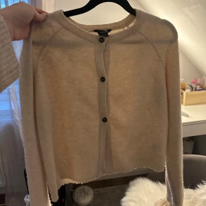 Beige kofta från Lindex med knappar - Supermjuk beige kofta från Lindex med rund halsringning och svarta knappar framtill. Långärmad och ribbade detaljer vid ärmslut och knappslå. Perfekt att slänga över en topp när det är kyligt ute.