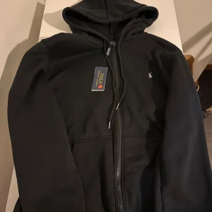 Svart hoodie från Polo Ralph Lauren - Svart hoodie från Polo Ralph Lauren med dragkedja och huva. Klassisk design med broderad logga på bröstet och praktiska fickor framtill. Tillverkad i mjuk bomull för en skön känsla. Perfekt för en chill och stilren look.