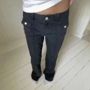 Svarta lågmidjade bootcut jeans - Säljer ett par svarta jeans med låg midja och bootcut-ben. Byxorna har framfickor med lock och silverfärgade knappar som ger en cool vibe. Materialet är klassisk jeans och passformen är normal. Perfekta för dig som gillar retrostil och vill sticka ut.