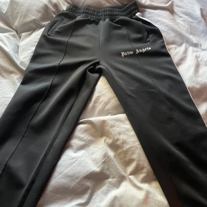 Svarta mjukisbyxor från Palm Angels - Svarta mjukisbyxor från Palm Angels med vit logotyp på vänster ben. Byxorna har elastisk midja och sidofickor, samt vita ränder längs sidorna. Materialet är mjukt och stretchigt, perfekt för en chill och sportig look.