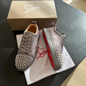 Gråa Christian Louboutin sneakers med nitar - Säljer ett par gråa sneakers från Christian Louboutin i mocka med ikonisk röd sula och silverfärgade nitar på tån. Skorna har snörning och en clean, lyxig vibe. Kommer med originalkartong och dustbag.