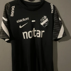 Svart AIK fotbollströja Nike - Säljer en svart AIK fotbollströja från Nike med klubbmärke på bröstet och flera sponsorer tryckta i vitt. Tröjan har korta ärmar med gråsvarta mönster och vita detaljer. Materialet är lätt och andas, perfekt skick med ett nupris på 999kr.