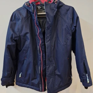 Blå skidjacka från Neomondo, junior - Snygg marinblå skidjacka från Neomondo med huva och dragkedja. Jackan har rosa detaljer på insidan och vid dragkedjan, samt praktiska fickor och justerbara muddar. Perfekt för blåsiga dagar och utomhusaktiviteter.