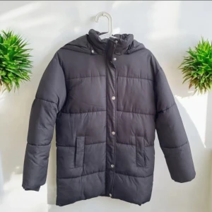 Svart pufferjacka med huva storlek 152 - Säljer en svart pufferjacka med huva och raka linjer. Jackan har dragkedja och tryckknappar framtill samt två stora fickor på sidorna. Perfekt för kalla dagar och enkel att matcha med olika outfits. Finns en reva som sluta ihop på baksidan , se bild 