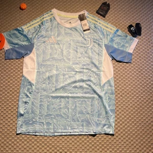 Juventus Yildiz 10 Adidas fotbollströja - Juventus fotbollströja från Adidas med Yildiz och nummer 10 på ryggen. Tröjan är ljusblå med vitt och har ett unikt abstrakt mönster, samt gula ränder på axlarna. Kortärmad modell i lätt och ventilerande Aeroready-material, perfekt för match eller träning.