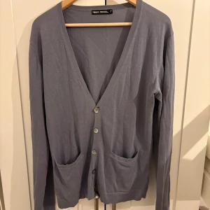 Ljusgrå kofta från Filippa K - Snygg ljusgrå kofta från Filippa K med v-ringning och knäppning framtill. Den har två fickor och är tillverkad i ett tunt, mjukt material som passar perfekt för lager-på-lager. Enkel och stilren design som funkar året runt.