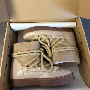 Beige boots från Inuikii med snörning - Snygga beige boots från Inuikii med grov snörning och platt brun sula. Utsidan är i mocka och skinn, insidan är fodrad med mjuk päls för extra värme. Perfekt för dig som vill ha både stil och komfort under kalla dagar.