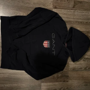 Svart hoodie från GANT med logga - Svart hoodie från GANT med klassisk logga och sköld på bröstet i rött, vitt och blått. Tröjan har huva, långa ärmar och en bekväm passform. Perfekt för dig som gillar stilrena plagg. Den är i storlek 170cm 15 år som är ca S i vuxenstorlek.