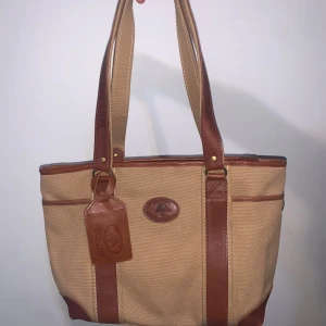 Beige handväska från Melvill & Moon - Snygg handväska i beige canvas med bruna skinn-detaljer från Melvill & Moon. Väskan har långa axelband, flera fack och ett randigt innerfoder i vit och ränder färger. Perfekt för dig som vill ha en stilren och praktisk väska med safari-vibes. Passar bäst kvinnor.