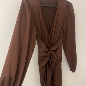 Brun omlottklänning från Gina Tricot - Snygg brun kort omlottklänning från Gina Tricot med långa ärmar och knytning i midjan. Klänningen har v-ringning och ballongärmar som ger en trendig siluett. Perfekt för dig som gillar stilrena och moderna plagg med en twist.