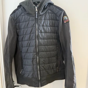 Svart quiltad jacka Parajumpers Y-L - Svart quiltad jacka från Parajumpers med huva och dragkedja framtill. Jackan har fleecepartier på ärmar och sidor, samt ett snyggt emblem på axeln. Perfekt för dig som gillar sportig och clean stil. Flera fickor med dragkedja och coola detaljer.