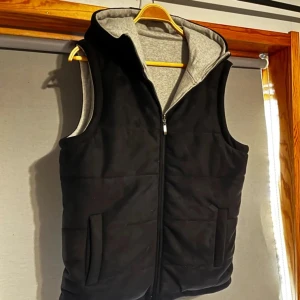 Svart och grå vändbar väst med huva - Snygg och praktisk vändbar väst med huva i svart och grått. Västen har dragkedja och är perfekt för både höst och vår. Den är tillverkad i ett mjukt syntetmaterial och har en normal passform.