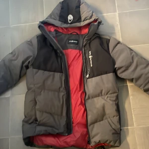 Sail racing jacka - Säljer en grå och svart pufferjacka från Sail Racing med röd insida. Jackan har huva, dragkedja framtill och flera praktiska fickor med dragkedja. Perfekt för kalla dagar och har en sportig vibe.
