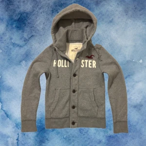 Grå Vintage Hollister Kofta/Cardigan Med Knappar - Väldigt sällsynt och vintage Hollister kofta/caridgan | Tröjan har stora knappar framtill, dragsko i huvan och två fickor | Perfekt Skick | Längd ≈ 61cm, Bredd Armhåla - Amrhåla ≈ 50cm  . | Storlek S! Modell 175, 68kg