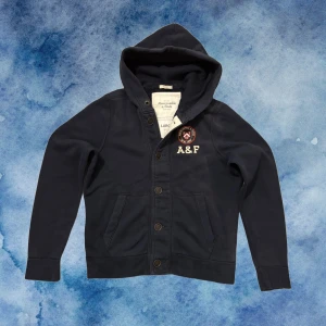 Vintage Abercrombie & Fitch Kofta/Cardigan Med Knappar - Säljer denna vintage och sällsynta Abercrombie & Fitch Cardigan med knappar | Storlek L men passar också M | Defekter: Urtvättad, snörre fattas samt ett liter hål över hålet för snörret | Längd ≈ 65cm , Bredd Armhåla - Armhåla ≈ 57cm