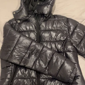 Svart pufferjacka med huva - Säljer en svart pufferjacka med huva och dragkedja framtill. Jackan har en glansig finish och är vadderad i breda kanaler för extra värme. Perfekt för kalla dagar och har en modern, sportig look. Har ett hål i fickan som går att fixa med nål och tråd, fickan går att stänga ändå. När fickan är stängd och jackan är på så syns inte hålet!