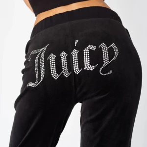 Svarta Juicy Couture mjukisbyxor - Svarta mjukisbyxor från Juicy Couture med glittrande strassdetaljer som bildar texten 'Juicy' över baken. Byxorna har en loose passform och är tillverkade i mjuk sammet för extra komfort. Perfekt för en chill och trendig look.