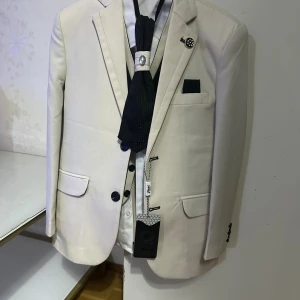 Beige kostym med svart väst och slips - Snygg beige kostym från Hikko med svart väst och matchande svart slips med dekorativ brosch. Kavajen har klassisk passform, svarta knappar och fickdetaljer. Västen har kontrasterande beige kant och knappar. Perfekt för dig som vill sticka ut med stilren look.