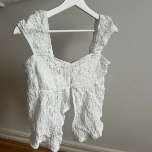 Vit ärmlös linnetopp med knappar - Säljer en vit linnetopp med breda axelband och snyggt rynkad struktur. Toppen har knappar framtill och är ärmlös, perfekt för varma dagar. Materialet känns lätt och luftigt, och den har en cool, lite skrynklig look som gör den unik.