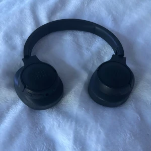 JBL trådlösa over-ear hörlurar - Snygga svarta JBL over-ear hörlurar med trådlös anslutning. Mjuka öronkuddar och tydliga L/R-markeringar. Inbyggda knappar för enkel kontroll. Hörlurarna ser ut att vara i gott skick utan synligt slitage eller defekter.