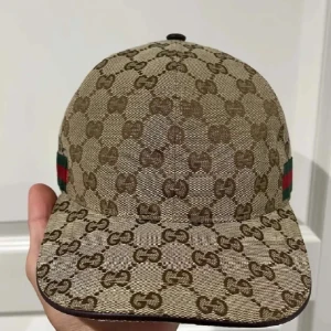 Gucci beige keps med GG-mönster - Snygg beige keps från Gucci med klassiskt GG-monogram över hela kepsen. På sidan finns en grön och röd rand som ger extra lyxig känsla. Justerbar rem baktill i brunt och insida i mörkbrunt tyg. Perfekt för dig som vill sticka ut med designerstil.
