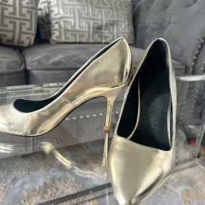 Guldiga pumps med spetsig tå från Ellos - Snygga pumps i metallic guld från Ellos med spetsig tå och smal klack. Skorna har en slät yta i syntetmaterial och svart innersula. Perfekt för dig som vill sticka ut med en glammig look och gillar eleganta detaljer.