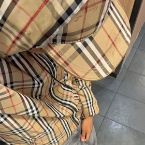 Burberry rutig vindjacka med huva - Burberry vindjacka med ikoniskt beige, svart och rött rutmönster. Jackan har huva med snörning, silverfärgad dragkedja och svart yttertyg. Insidan och huvan har klassiskt Burberry-mönster. Perfekt för dig som vill ha en clean och lyxig streetstil. Ps finns en liten skada på vänster axel