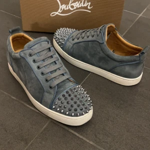 Christian Louboutin skor - Säljer ett par grymma blå sneakers från Christian Louboutin i mocka med coola silverfärgade nitar på tån. Skorna har snörning, vit platt sula och den ikoniska röda undersidan. Insidan är beige och de har en snygg rund tå. Perfekt för dig som vill sticka ut med din stil. Bra skick 9/10, Priset är ej hugget i sten