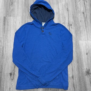 Blå hoodie från Abercrombie & Fitch - Snygg blå hoodie från Abercrombie & Fitch i storlek M. Tröjan har huva, knappar vid halsen och ett diskret logotryck på bröstet. Tillverkad i mjuk bomull med långärmad passform och känguruficka framtill. Perfekt för chill dagar.