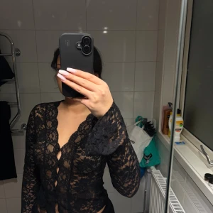 Svart spetsblus med v-ringning - Säljer en svart blus i genombruten spets med v-ringad front och långa ärmar. Blusen har ett croppat snitt och vackra spetsdetaljer över hela plagget. Perfekt för dig som gillar en edgy och feminin stil.