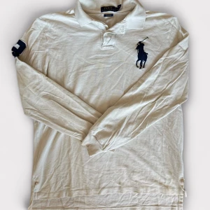 Vit långärmad pikétröja Polo Ralph Lauren - Vit långärmad pikétröja från Polo Ralph Lauren med klassisk krage, stor mörkblå broderad polospelare på bröstet och siffra 3 på ärmen. Tröjan är i bomull och har en normal passform. Perfekt för en clean och sportig stil.