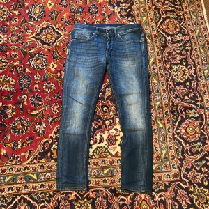 Blå skinny jeans från Dondup George - Säljer ett par blå jeans från Dondup, modell George Skinny Fit. Jeansen har klassisk femficksdesign, snygga slitningar och smal passform som sitter tajt längs benen. Materialet är jeans med orangea sömmar och cool tvättad look.