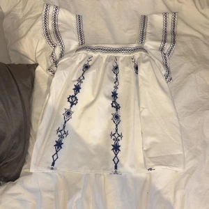 Vit blus med blå broderier - Säljer en söt vit blus med blå broderade detaljer och volangärmar. Blusen har raka axelband och vackra blommönster framtill. Perfekt för dig som gillar boho-stil och vill ha något unikt i garderoben. Perfekt till Grekland vibes och sommar romanser.