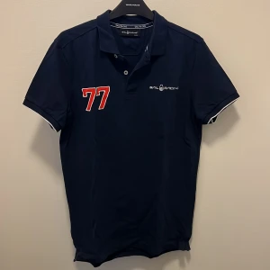 Marinblå pikétröja Sail Racing - Snygg marinblå pikétröja från Sail Racing med vit logga och röd 77-tryck på bröstet. Klassisk krage, korta ärmar med vit kant och knappar framtill. Perfekt för dig som gillar sportig och stilren look.