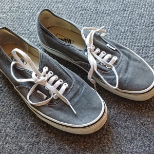 Gråa Vans sneakers med vit sula - Klassiska Vans sneakers i grå canvas med vita snören och vit sula. Skorna har låg profil, rund tå och den ikoniska gummisulan med rutmönster. Perfekta för en avslappnad streetstil och passar till det mesta.